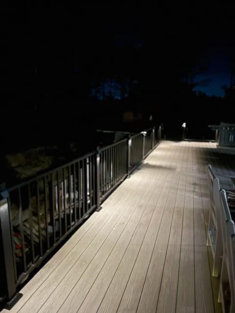 Long composite deck corridor at night with post-cap lighting, black metal railing, and wooded surroundings