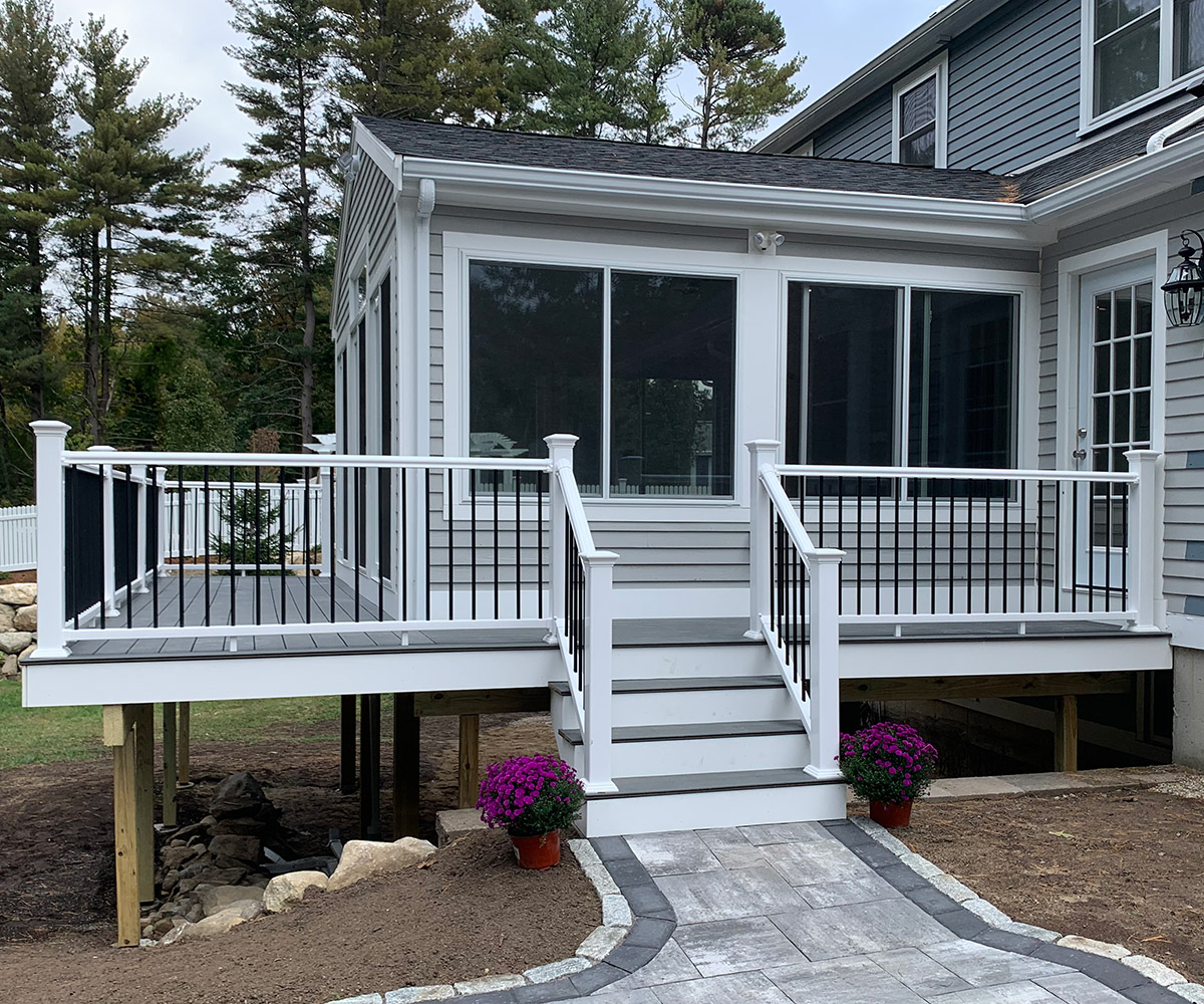 A newly constructed, elevated deck with black and white railings is attached to a gray house. Steps lead down to a stone pathway, and purple flowers in pots sit at the bottom of the stairs.