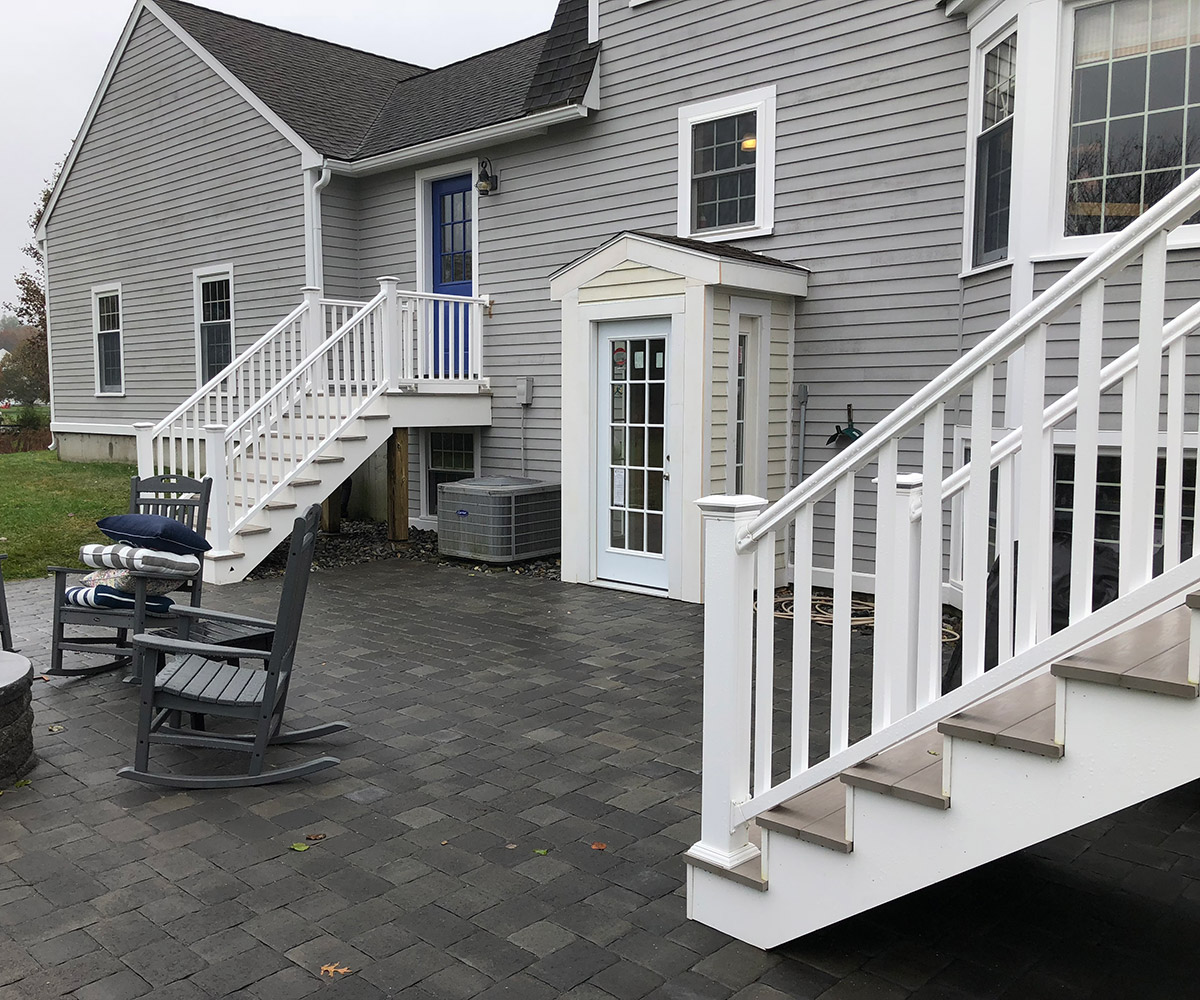 A two-story gray house with white trim and a small upper-level balcony with a white railing. In front, there is a paved patio area with dark stone tiles, featuring a set of stairs leading up to the balcony. To the left, there is an outdoor rocking chair.