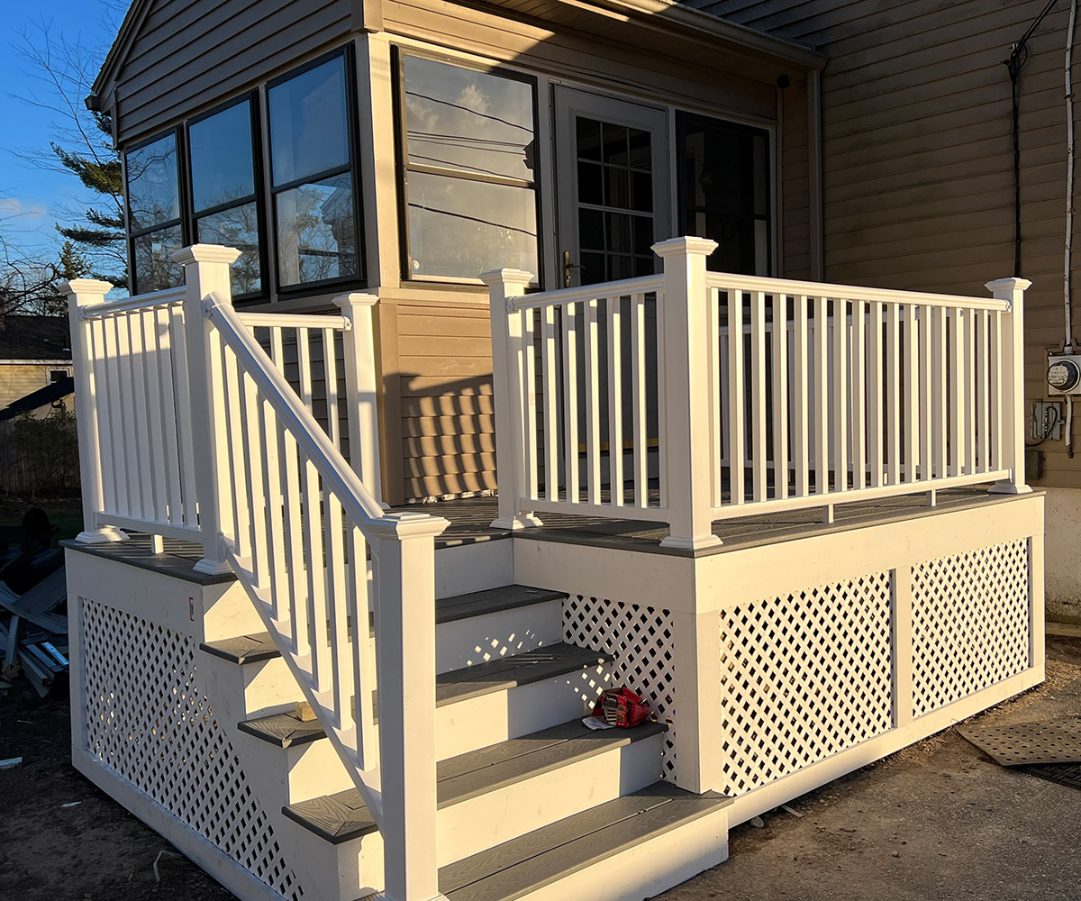 A recently renovated porch with white railings and stairs. The porch features latticework on the sides and gray wooden steps leading up to a glass door. The structure is attached to a house with tan siding, and the scene is lit by late afternoon sunlight.