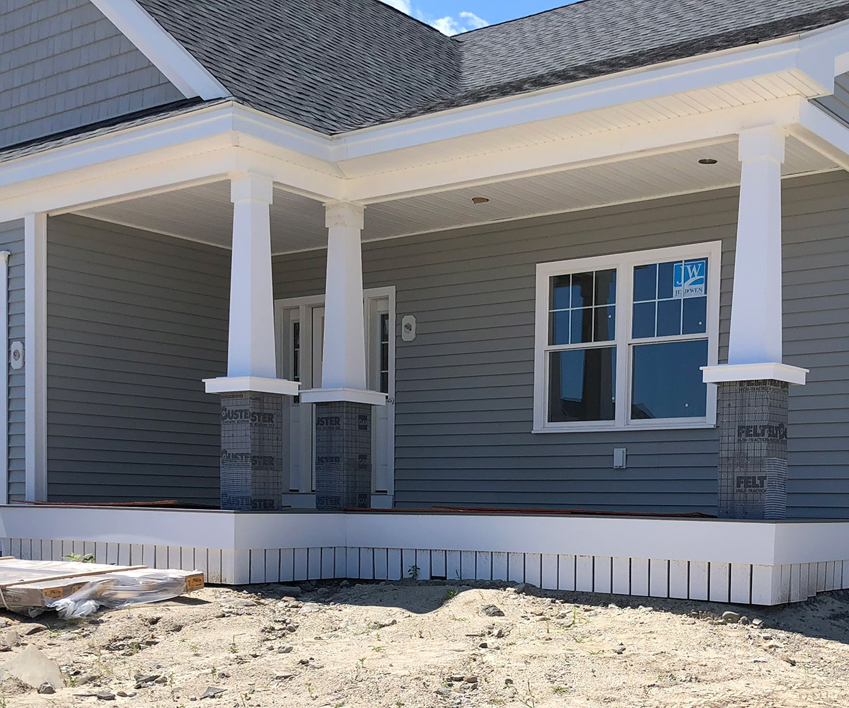 A newly constructed porch with white trim and large, tapered columns wrapped in protective covering. The house has gray siding and white-framed windows. The porch floor is unfinished, and the surrounding ground is bare, indicating ongoing construction. The roof is covered with gray shingles, and the sky is clear and blue.