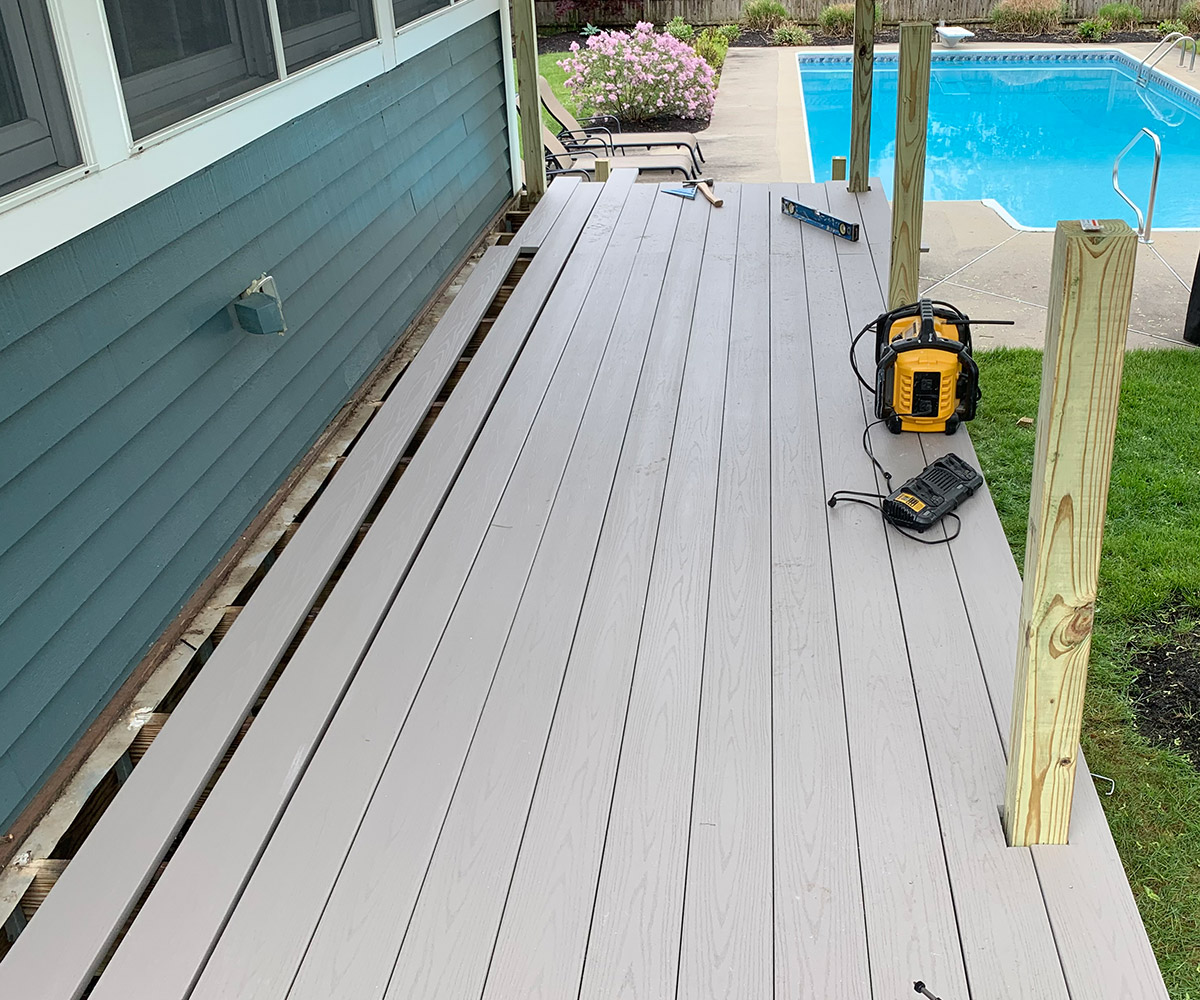 A deck under construction with gray composite decking boards being installed. The deck is attached to a house with teal siding and features an electric tool and level on the unfinished surface. The area around the deck includes a grassy yard and a swimming pool, with blooming pink flowers visible in the background.