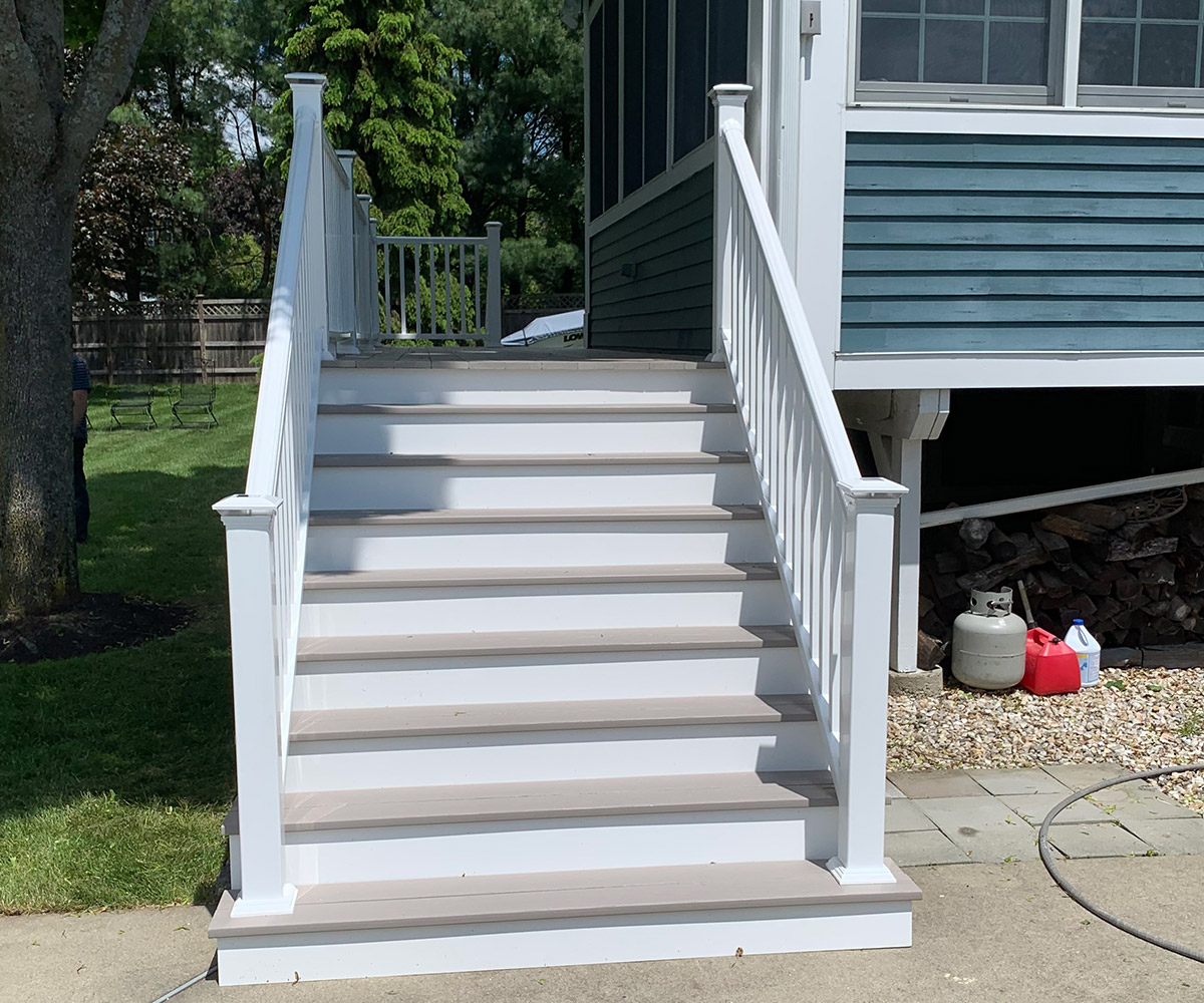 A set of new composite board stairs leading up to a blue house with white trim. The stairs are flanked by white railings with classic design posts, and there is a neatly trimmed green lawn on either side. The sky is clear and blue, indicating a sunny day.