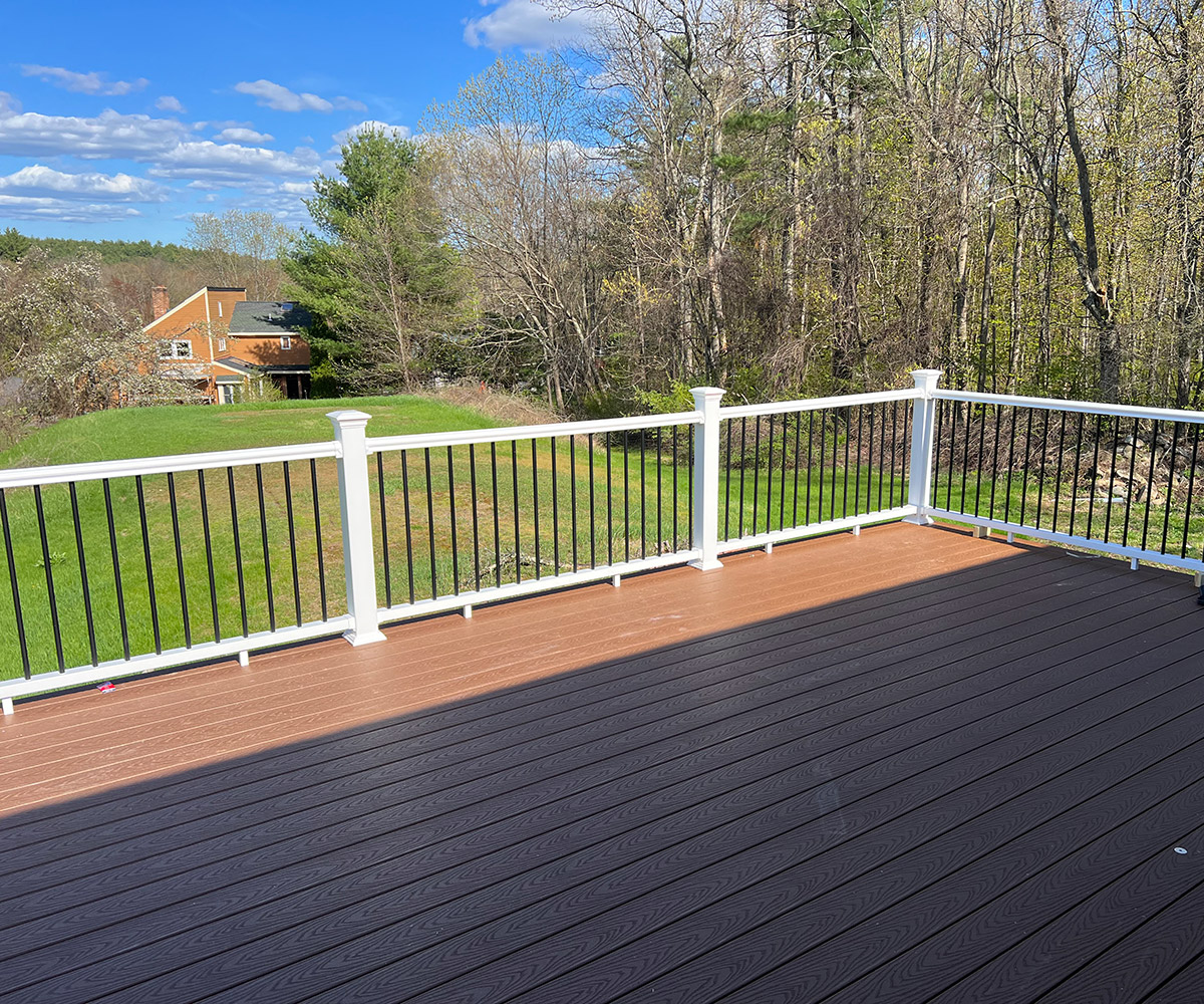 A spacious outdoor deck with dark brown flooring and white railings. The deck overlooks a backyard with green grass, trees with early spring foliage, and a house in the distance under a clear blue sky with scattered clouds.