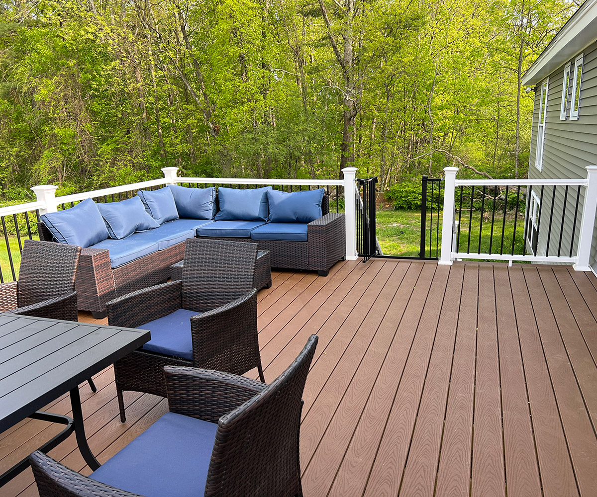 An outdoor patio area with dark brown composite decking. The space is furnished with a modern wicker sectional sofa adorned with blue cushions and a matching wicker coffee table. A black dining table with six chairs is also present. The patio is attached to a light grey house and is surrounded by white railings, overlooking lush green trees in the background, indicating a suburban setting.