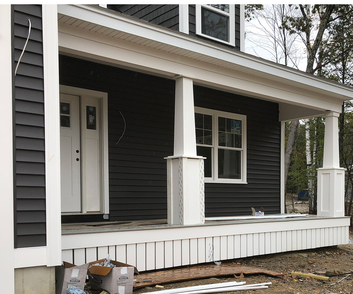 A two-story house with dark gray siding and white trim. The house features a covered front porch with white columns and railings. A white front door is flanked by sidelights and a single window to the right.