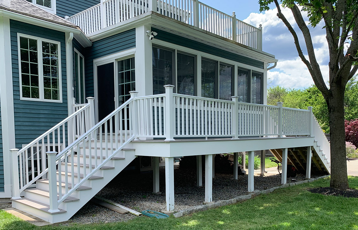 An exterior view of a house with a two-level deck. The upper deck is enclosed with large windows and has a white railing. There is a staircase leading down from the upper deck to the lower deck, which is also supported by white posts and has a matching white railing. The house exterior is blue with white trim around the windows and doors. The area under the deck is open, revealing a gravel-covered ground. A tree and a lawn are visible in the foreground.