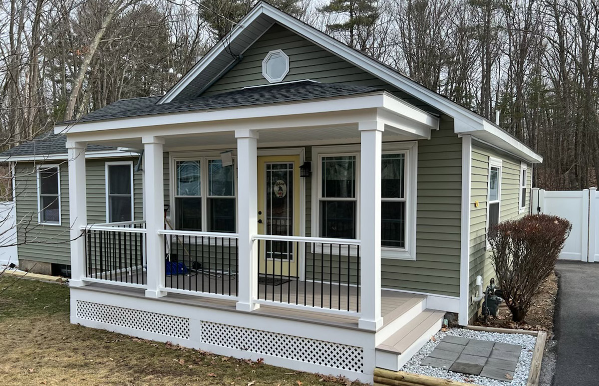 A small house with a covered front porch. The house exterior is green with white trim, and the porch has white columns supporting a roof. The porch railing is white with black balusters, and the base of the porch has white latticework. The front door is yellow with a decorative oval window. There are three windows on the front of the house, and the lawn around the house appears to be well-maintained with a mix of grass and a small gravel area. Trees are visible in the background.