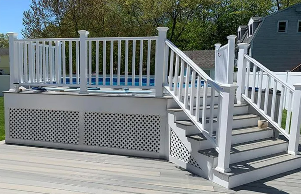 A raised deck with a white railing and steps leading up to an above-ground pool. The base of the deck is enclosed with white lattice panels. The steps have white railings that match the deck, and the area surrounding the deck appears to be a well-maintained yard with grass and some trees in the background. There is a house and a white fence visible in the distance.